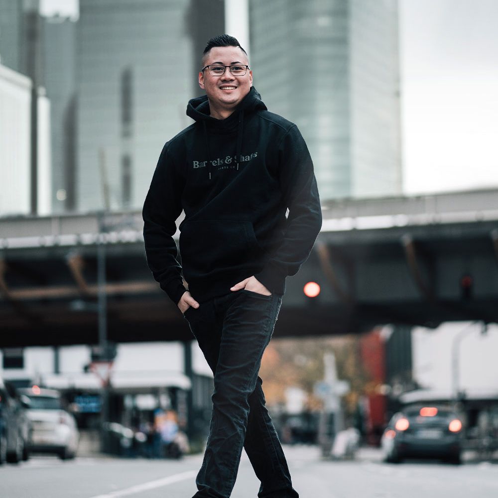 Barrels and Shafts Hoodie - Black Eine Person trägt einen schwarzen "Barrels and Shafts" Hoodie und steht entspannt auf einer Straße. Im Hintergrund sind unscharfe Gebäude und Autos zu sehen.