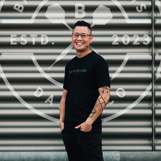 Barrels and Shafts T-Shirt - Black Ein Mann trägt ein schwarzes "Barrels and Shafts"-T-Shirt und steht vor einer metallenen Wand mit großem Logo. Er lächelt und hat markante Tattoos am rechten Arm.