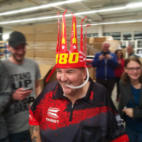 9026-McDart-180-er-Tr-tenkrone-4z2QvH75NbVwnP Ein lachender Mann trägt die "McDart 180-er Trötenkrone" auf dem Kopf. Um ihn herum stehen weitere Menschen und lachen ebenfalls.